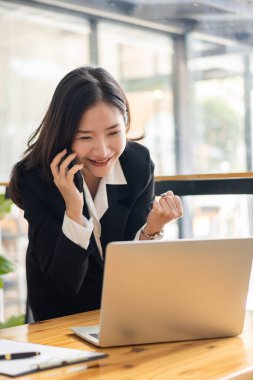 Asyalı kadın ofis çalışanı Brand, ve analist, modern bir ofiste dizüstü bilgisayarın önünde oturuyor. İyi haberle sevinin, anlaşmayı tamamlayın ve gülümseyin..