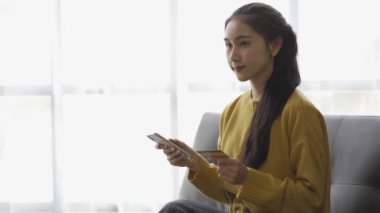 Asyalı bir kadın elinde kredi kartıyla akıllı telefonuyla alışveriş yapıyor ve evinde, kanepede ödeme yapıyor.