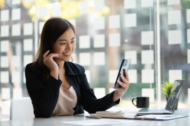 Başarılı Asyalı iş kadını elinde akıllı bir telefonla ofisteki masada dizüstü bilgisayarın finansal grafik verileri üzerinde çalışıyor..