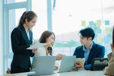 Üst düzey bir ofisteki çok ırklı genç bir iş ekibi dizüstü bilgisayarda oturup mutlu bir gülümsemeyle evrak işleriyle uğraşıyor. Asyalı iş adamları grubu modern ofis tasarım ve konsept planlamada toplanıyor.