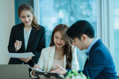 Üst düzey bir ofisteki çok ırklı genç bir iş ekibi dizüstü bilgisayarda oturup mutlu bir gülümsemeyle evrak işleriyle uğraşıyor. Asyalı iş adamları grubu modern ofis tasarım ve konsept planlamada toplanıyor.