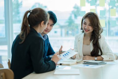 Üst düzey bir ofisteki çok ırklı genç bir iş ekibi dizüstü bilgisayarda oturup mutlu bir gülümsemeyle evrak işleriyle uğraşıyor. Asyalı iş adamları grubu modern ofis tasarım ve konsept planlamada toplanıyor.
