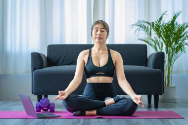 Evde yoga yapan güzel bir Asyalı kadın. Oturma odasında omuzlarına sağlıklı bir gülümseme geriliyor. Geniş bir pencerenin yanında tüm vücut portresi, spor ve sağlıklı yaşam tarzı kavramları.