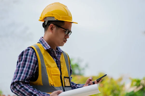 A young professional engineer in a protective helmet and blueprints at a young man\'s house building construction site with a smartphone checking work on documents on the construction site.
