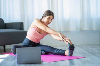 Genç ve güzel bir Asyalı kadın dizüstü bilgisayarda egzersiz yaparken kollarını uzatıyor. Genç bir kadın oturma odasında yoga pozlarını ısıtıyor. sağlık hizmeti yaşam tarzı iyi sağlık konsepti