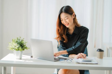 Gülümseyen mutlu Asyalı kadın masadaki bir sandalyede otururken dizüstü bilgisayar teknolojisini kullanarak rahatlıyor. Evde çalışan yaratıcı kız. İnternette öğrenim görme ve çalışma kavramı.