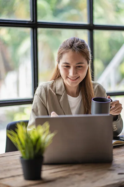 Gülümseyen mutlu Asyalı kadın masadaki bir sandalyede otururken dizüstü bilgisayar teknolojisini kullanarak rahatlıyor. Evde çalışan yaratıcı kız. İnternette öğrenim görme ve çalışma kavramı.