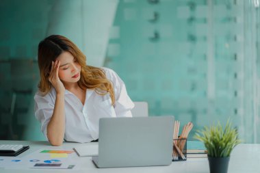 Asian businesswoman having a headache during work analyzing financial graphs with laptop at office desk