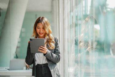 Ofiste cam duvarın yanında tabletle duran Asyalı iş kadını.