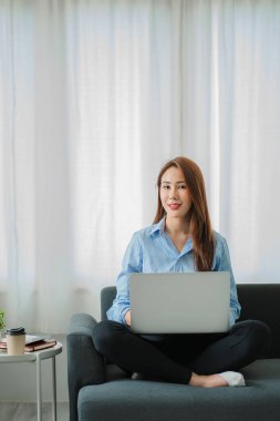 Güzel bir Asyalı kadının portresi kanepede oturuyor ve evdeki oturma odasında dizüstü bilgisayarıyla çalışıyor. Evden çalışıyorum. Yeni Normal ve Dostane Konsept.