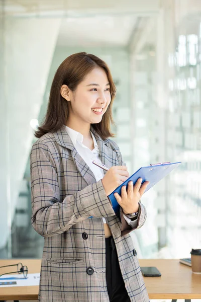 Attractive Asian woman holding a folder standing in front of a desk in a financial accounting concept office, vertical image.