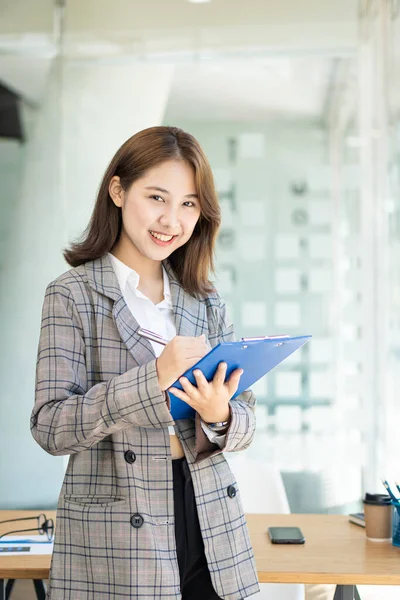 Attractive Asian woman holding a folder standing in front of a desk in a financial accounting concept office, vertical image.