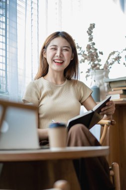 Asyalı bir kız masada elinde dizüstü bilgisayarla mutlu bir şekilde oturuyor ve masa başı konseptinde çalışmayı öğreniyor.
