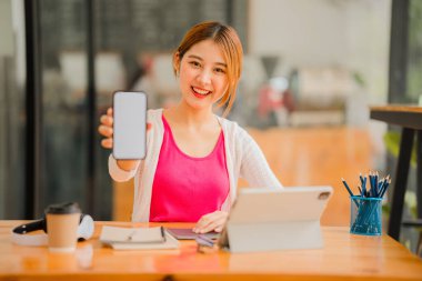 beautiful Asian girl A cute girl shows a big smartphone with a white blank screen in her hand. Sitting at a table with a tablet