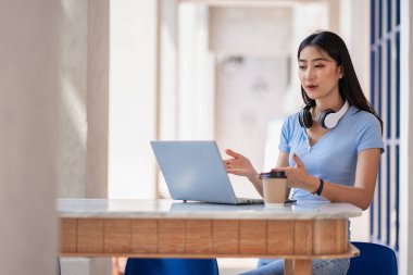 Şirin bir Asyalı öğrenci, öğretmeniyle çevrimiçi çalışmak için bilgisayarında çalışırken boynuna kulaklık takıyor. Kız çalışmaktan mutlu ve bir kafede ders dinliyor..