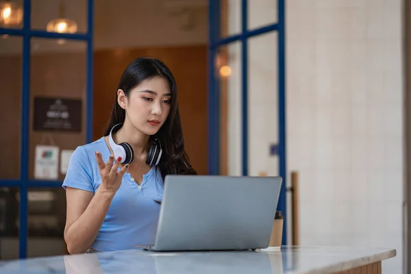 A cute Asian student hangs headphones around her neck while studying online on her laptop to study online with her teacher. the girl is happy to study and Listen to a lecture inside a coffee shop.