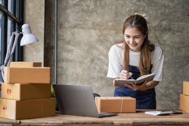 Küçük işletme kuruluşu KME, Asyalı bir bayan girişimcinin sahibi, paketleme kutularını hazırlamak için online siparişleri almak ve kontrol etmek için bir dizüstü bilgisayar kullanıyor. İnternette fikirler satmak, kargo paketleri.