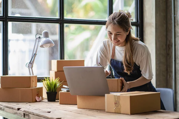 Small business start-up, SME, owner of an Asian female entrepreneur uses a laptop to receive and check online orders to prepare packing boxes. Selling ideas online, shipping parcels.