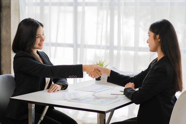Two smiling female business partners shaking hands in the office teamwork office girl to make a deal in a meeting to discuss financial graph and laptop computer