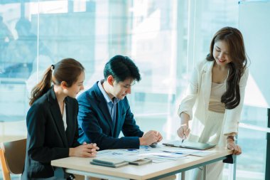 Genç Asyalı iş adamları finansal belgelerde dizüstü bilgisayarlarla mutlu bir şekilde çalışıyorlar. Bir grup iş adamı modern bir ofis tasarım ve konsept planlamada toplanıyor.