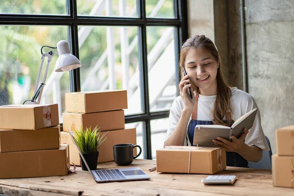 Küçük işletmeler, KME sahipleri, Asyalı bayan girişimciler kutular, dizüstü bilgisayarlar, akıllı telefonlar ile çalışıyorlar. KOBİ müşterilerine satmak üzere paket göndermek üzere kutuları paketlemeye hazırlanmak için çevrimiçi siparişi işaretleyin. Çevrimiçi iş fikirleri.