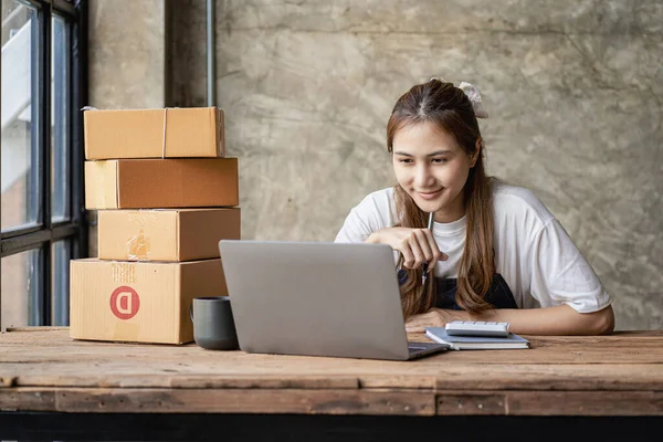 Küçük bir işletmenin portresi, KME sahibi, genç Asyalı girişimci, çalışıyor, kutuları açıyor, siparişleri kontrol ediyor. KME müşterilerine satılmak üzere paket teslimat kutularını çevrimiçi iş fikirlerine hazırlamak için