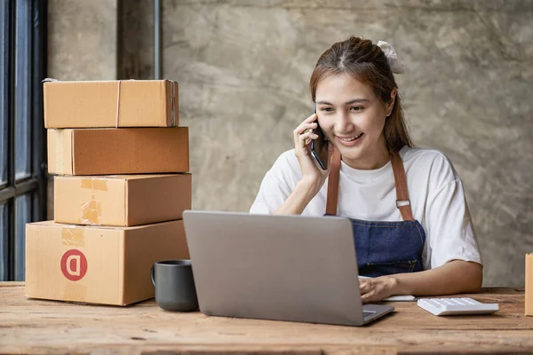 Small business startups, SME owners, Asian female entrepreneurs Work with boxes and laptops, smartphones. check order online To prepare to pack boxes to send parcels to sell to SME customers. Online business ideas.