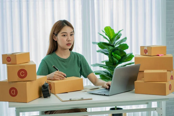 Küçük bir işletmenin portresi, KME sahibi, genç Asyalı girişimci, çalışıyor, kutuları açıyor, siparişleri kontrol ediyor. KME müşterilerine satılmak üzere paket teslimat kutularını çevrimiçi iş fikirlerine hazırlamak için