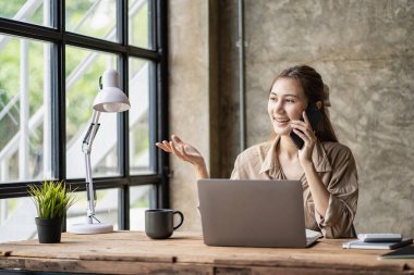 Business and communication concept, Asian woman working on laptop computer and reading smartphone on internet online at home, independent girl smiling using phone with social media.