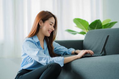 Asian woman sitting on sofa at home using digital tablet for work or online shopping. Touch screen freelance device for business or social media reading.
