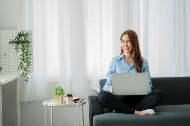 A happy Asian woman using a laptop computer sits on the sofa in the living room. work from home concept