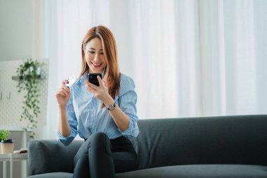 Asian woman in apartment holding device in hand Make an online purchase, enter credit card details. Type a letter on your phone and pay via internet banking.