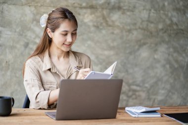 Smiling Asian woman working with laptop computer at her desk in modern home office. calculating financial expenses Make a note on the graph paper data sheet.