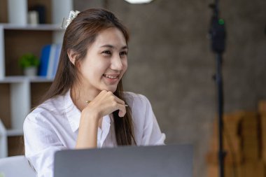 Smiling Asian woman working with laptop computer at her desk in modern home office. calculating financial expenses Make a note on the graph paper data sheet.
