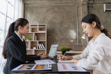 Two young Asian female accountants discuss working on investment projects and planning strategies using laptop computers in their office.