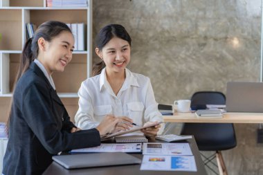 Two young Asian female accountants discuss working on investment projects and planning strategies using laptop computers in their office.