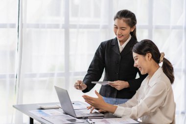 Two young Asian female accountants discuss working on investment projects and planning strategies using laptop computers in their office.