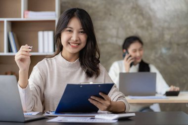 Two young Asian female accountants discuss working on investment projects and planning strategies using laptop computers in their office.