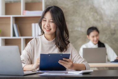 Two young Asian female accountants discuss working on investment projects and planning strategies using laptop computers in their office.