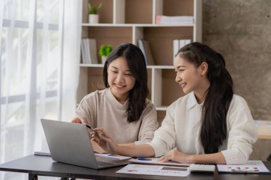Two young Asian female accountants discuss working on investment projects and planning strategies using laptop computers in their office.