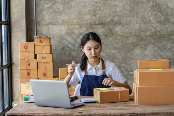 Starting a Successful Small Business Owner Independent Asian female saleswoman using laptop to check and Prepare parcel boxes to send to customers, SME entrepreneurs, online business ideas