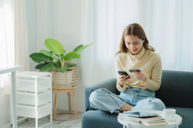 Asian female consumers with credit cards and smartphones sit on their sofas at home to do banking and mobile online shopping.