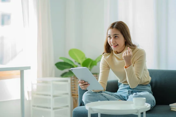 Asian woman holding a tablet on the sofa at home doing financial transactions and online shopping.