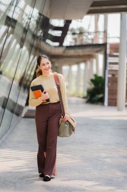 Asian woman holding a tablet and shoulder bag walking beside the educational concept building