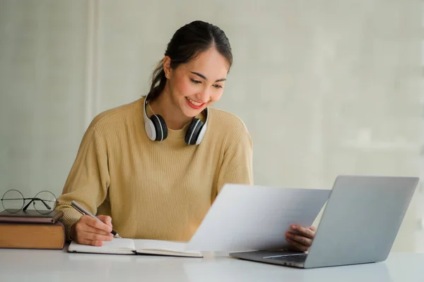 A young Asian student studying online using headphones and a laptop takes notes in a notebook sitting at her desk at home.