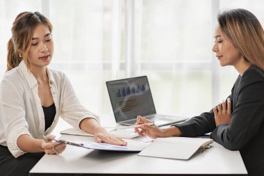 Two Asian business women working on a financial graph and a laptop.in the presentation and review of the business plan Joint financial marketing strategies in the meeting room with new employees in the office.