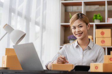 New business owner Asian woman working at home office Packaging on background SME entrepreneur online shopping or freelance work concept Young woman happy after new order from customer
