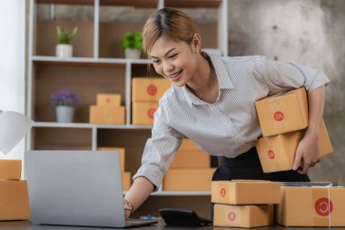New business owner Asian woman working at home office Packaging on background SME entrepreneur online shopping or freelance work concept Young woman happy after new order from customer