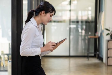 A young accountant stands with her laptop next to a glass wall in her office.