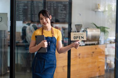 A beautiful Asian barista in an apron holds a plaque and stands in front of a cafe door with an open sign.